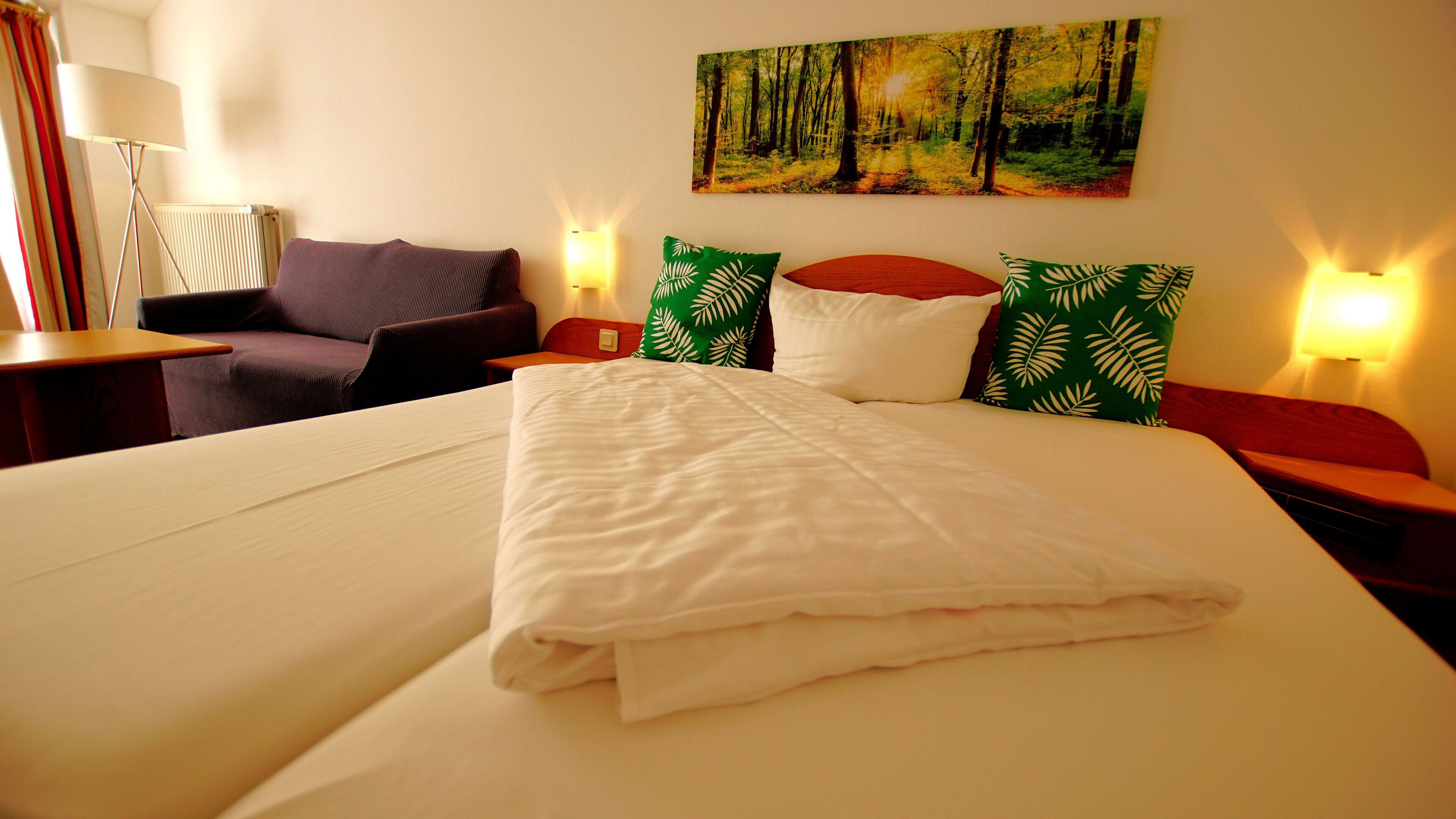 Una cama bien hecha con sábanas blancas, una almohada estampada en verde y blanco, y una almohada roja sobre un cabecero de madera. En la pared sobre la cama, hay un cuadro de un bosque.