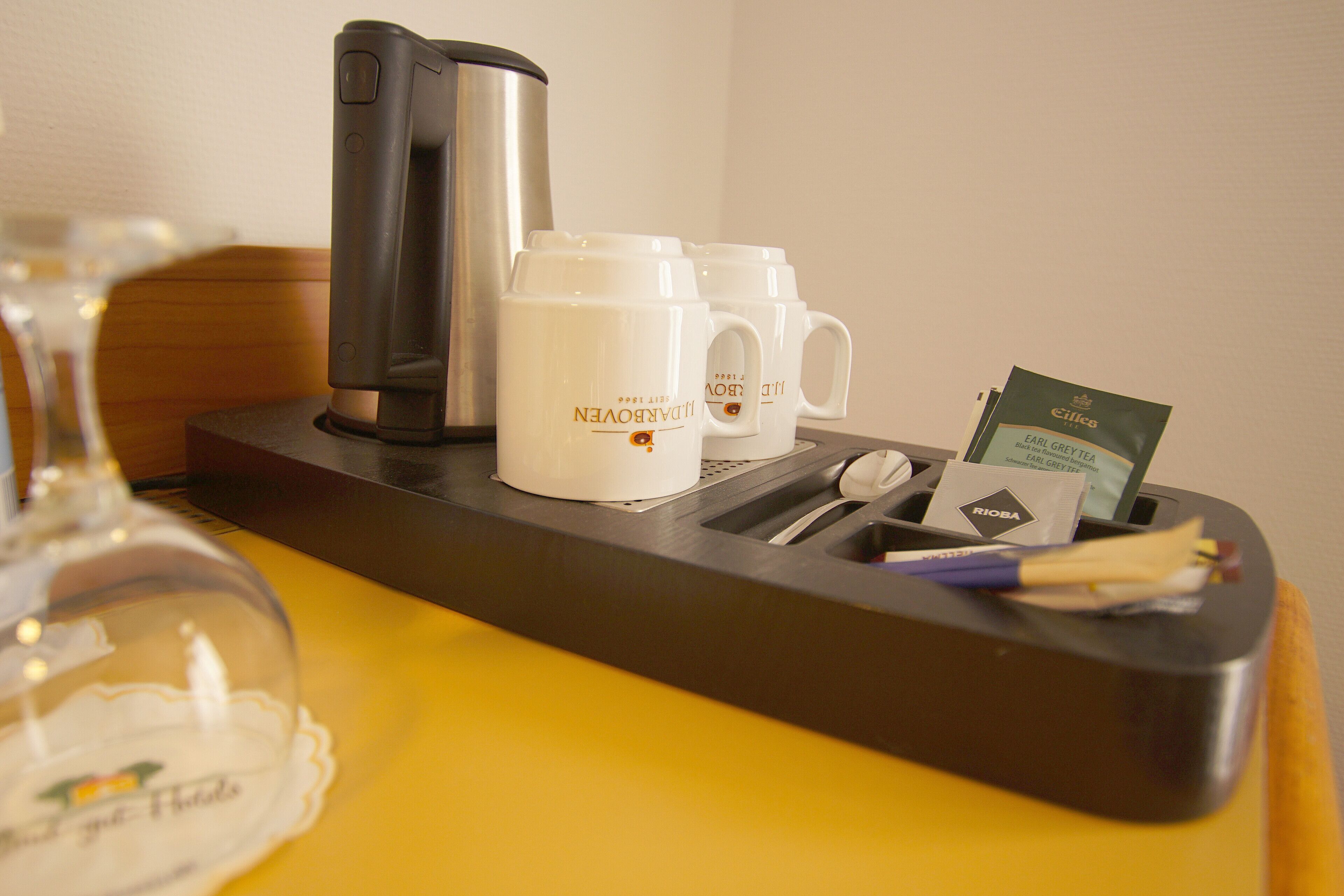 A hotel room's minibar with a coffee maker, two mugs, a glass, and a card holder.