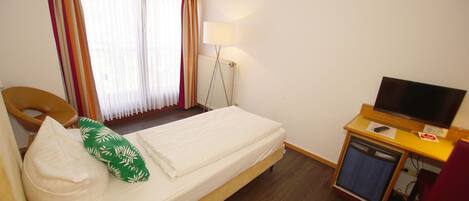 A single bed with a green pillow, a wooden chair, a desk with a television, and a window with curtains.