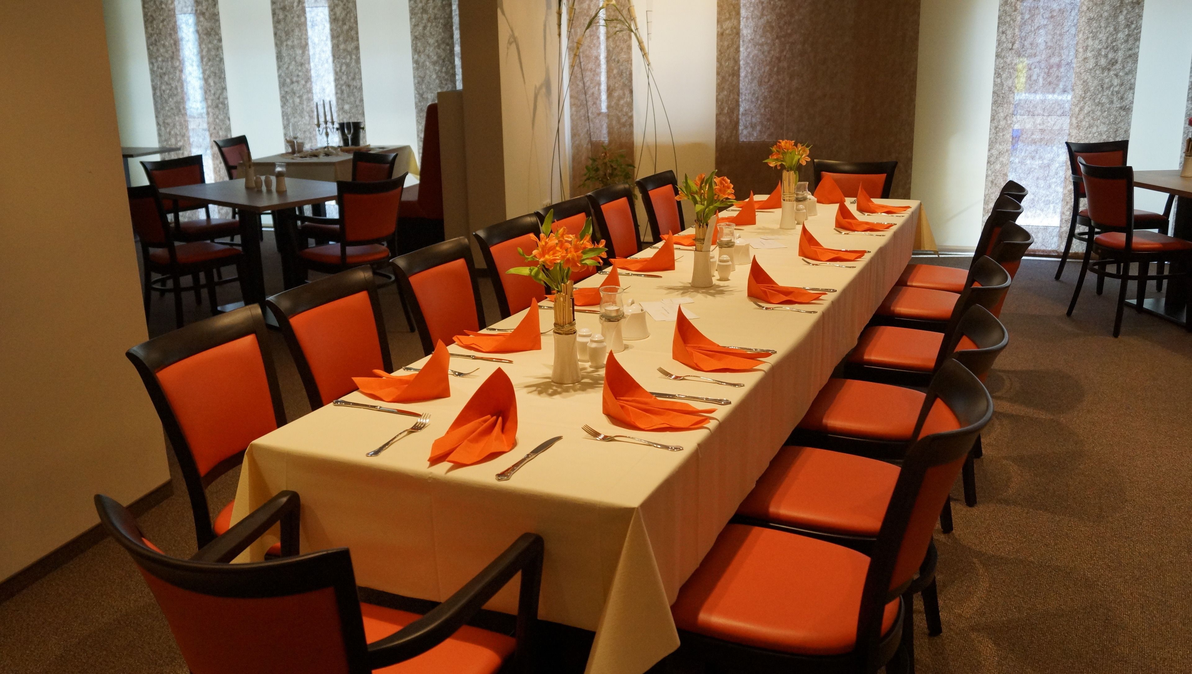A long dining table set with orange napkins, silverware, and vases of flowers.