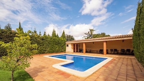 Una piscina con solárium empedrado, rodeada de vegetación y un edificio con terraza.
