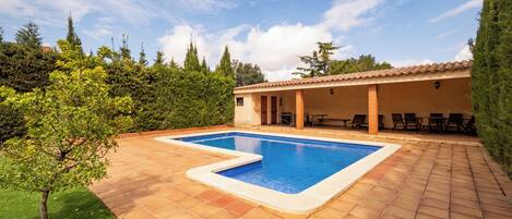 Una piscina con solárium empedrado, rodeada de vegetación y un edificio con terraza.