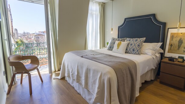 A bedroom with a large bed, a chair, a nightstand, and a view of the city through a window. - Bairro Alto Suites (Lisbon)