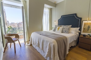 Een slaapkamer met een groot bed, een stoel, een nachtkastje en uitzicht op de stad door een raam.
