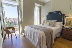 Un dormitorio con una cama grande, una silla, una mesita de noche y vistas a la ciudad a través de una ventana.