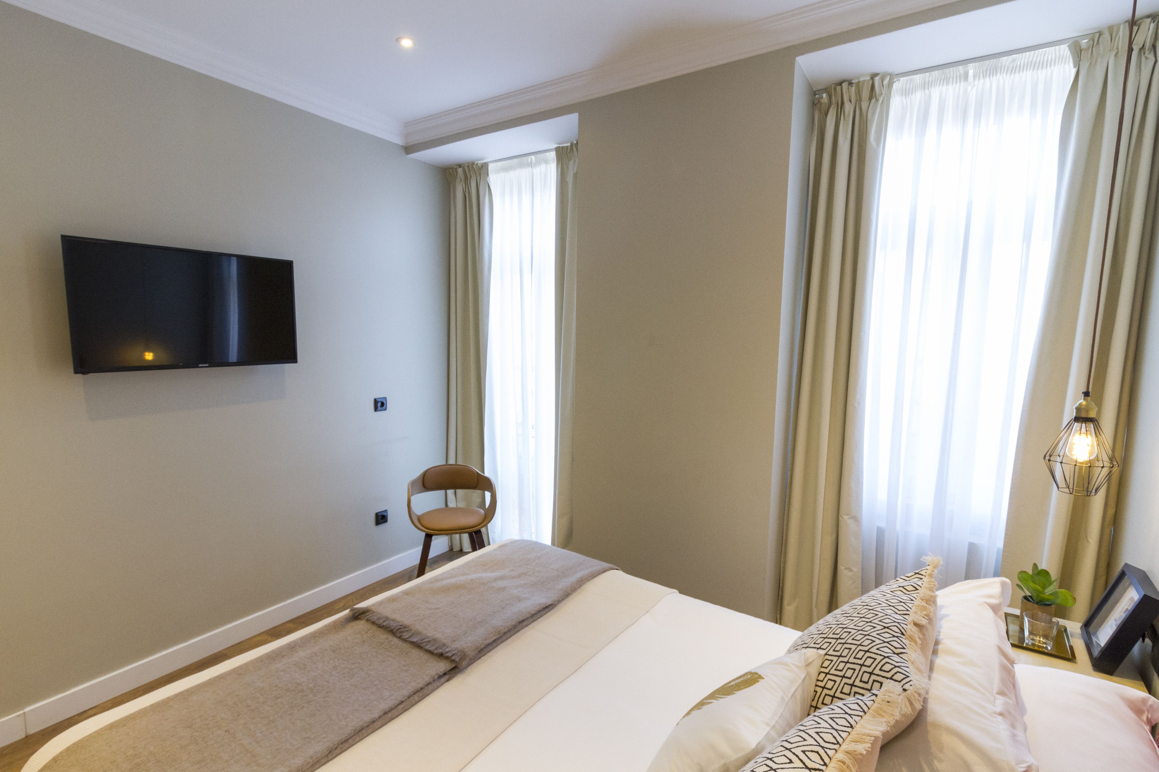 Un dormitori amb un llit, una cadira, un televisor fixat a la paret i una finestra amb cortines.