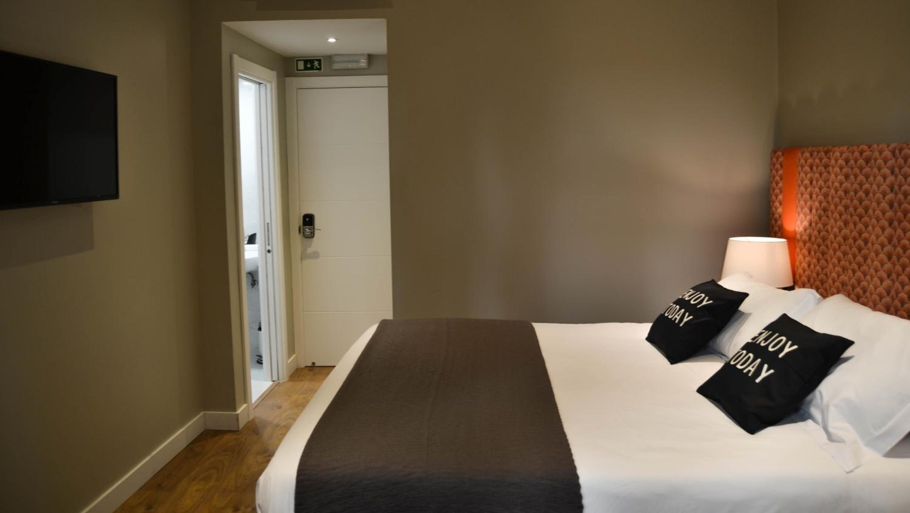 A hotel room with a bed, a TV mounted on the wall, a door, and a wall-mounted light.
