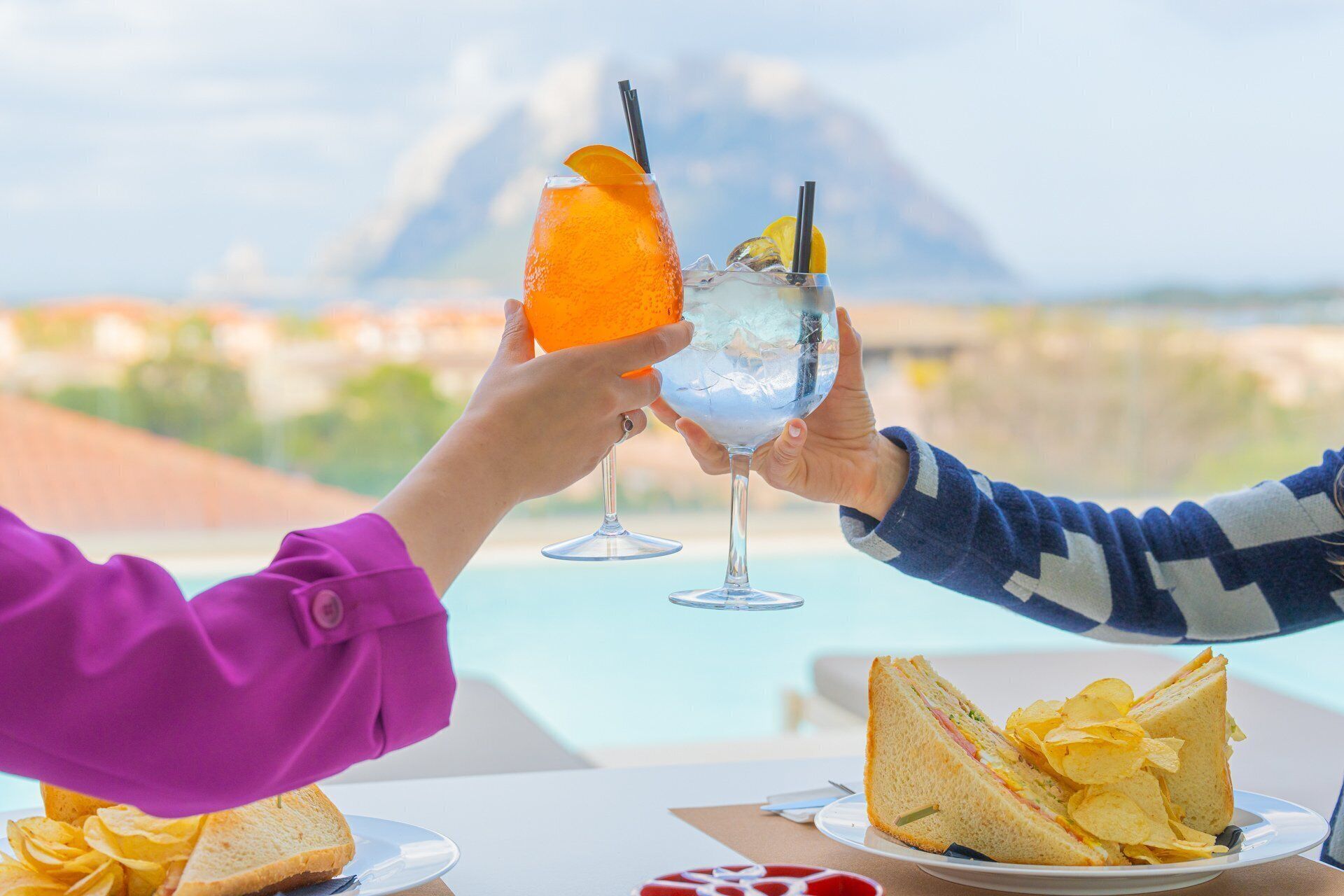 Duas pessoas brindando com bebidas, um sanduíche, batatas fritas e uma vista de montanhas ao fundo.