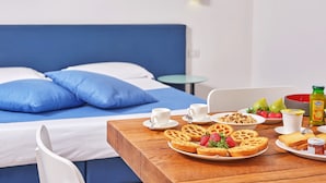 Een netjes opgemaakte ontbijttafel met wafels, aardbeien en sap in een hotelkamer met een blauw bed en witte muren.