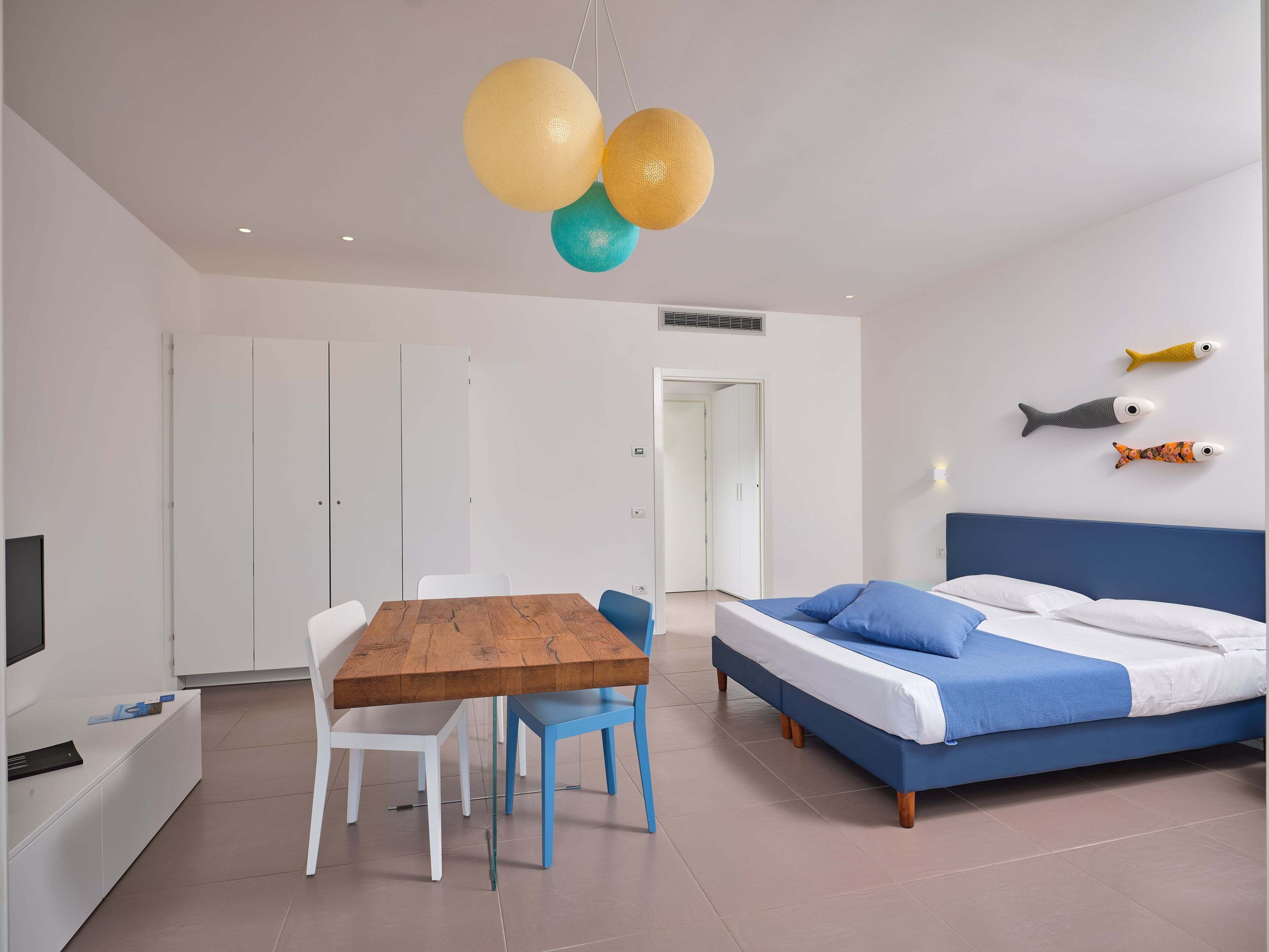 A modern bedroom with a wooden dining table, white chairs, a blue bed, and a wall-mounted fish decoration.
