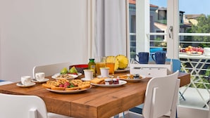 Een ontbijttafel met verschillende soorten eten en drinken, witte stoelen en een uitzicht op gebouwen door een raam.