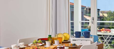 A breakfast table with various food items and drinks, white chairs, and a view of buildings through a window.