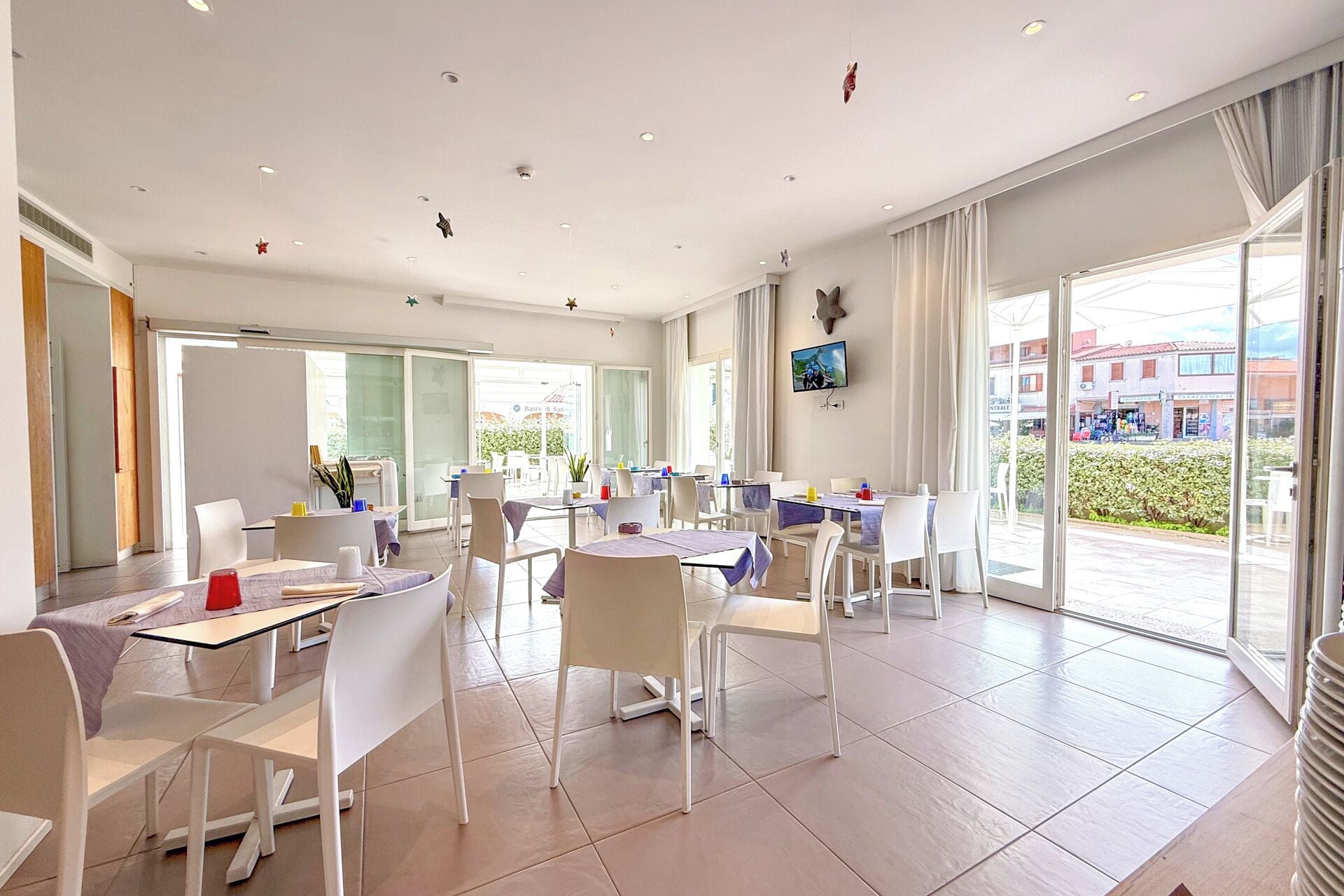 A modern dining area with tables and chairs, large windows, and a television on the wall.
