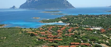 Vista aérea de una zona costera con una isla rocosa destacada al fondo, rodeada de aguas azules cristalinas. Se observan numerosos edificios con tejados rojos, probablemente viviendas o pequeños hoteles, dispersos por el paisaje.