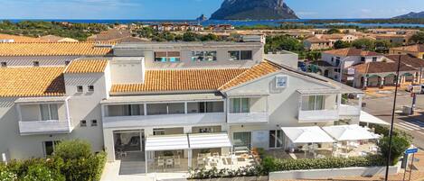 A modern hotel with a terrace, a view of the sea, and a prominent mountain in the background.