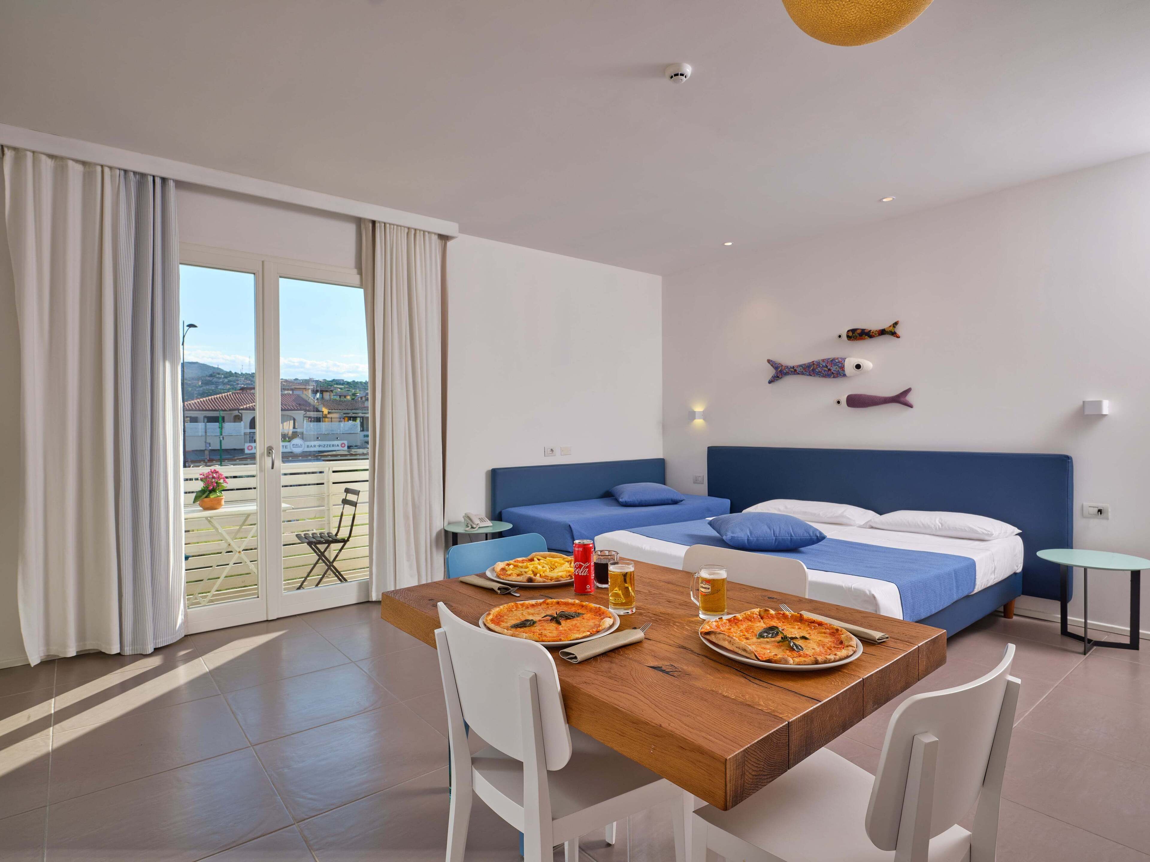 A modern hotel room with a balcony, a dining table with pizza, and a bed with blue bedding.