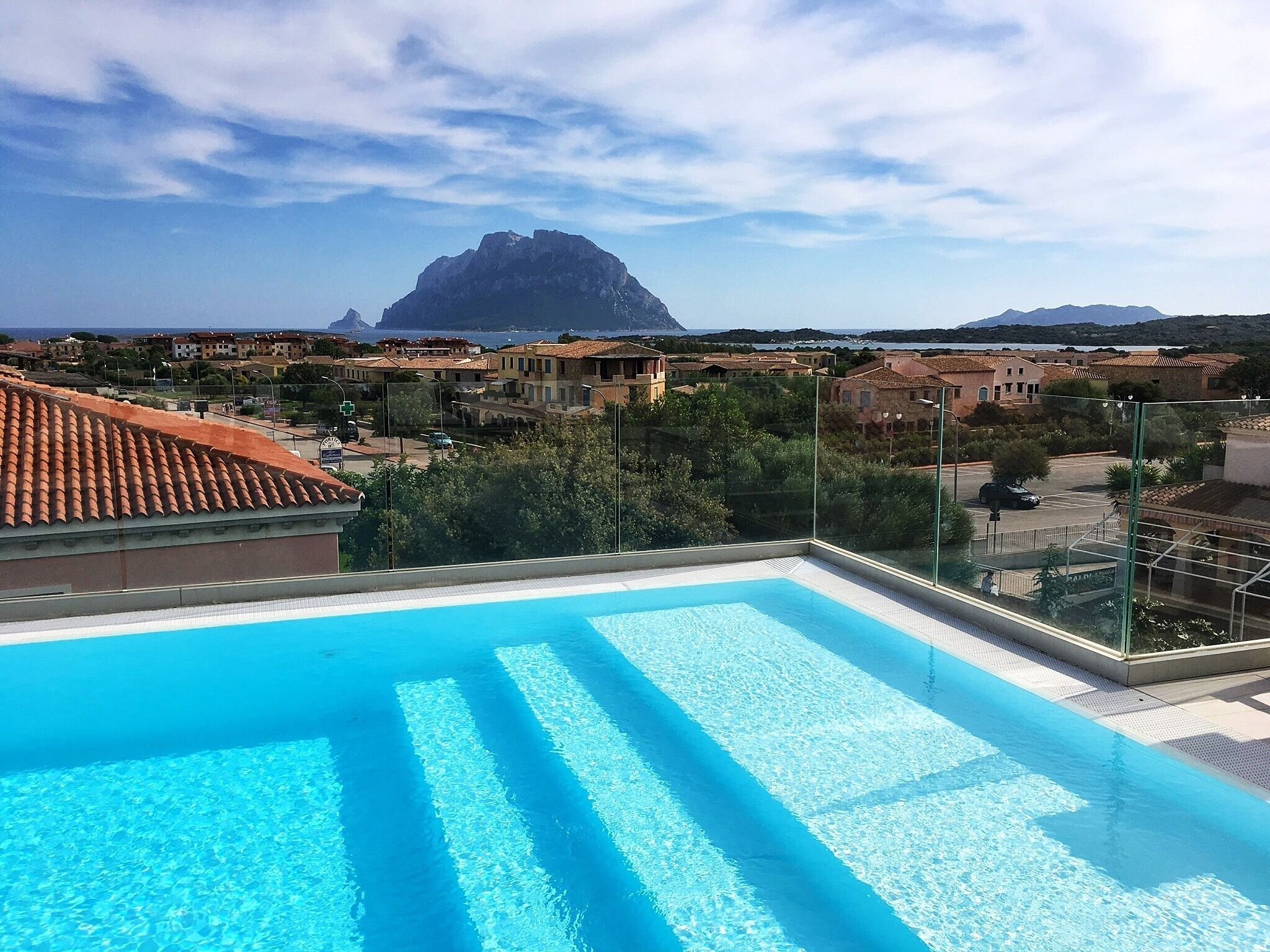 A swimming pool with clear blue water, surrounded by buildings with red-tiled roofs, and a mountain in the background.