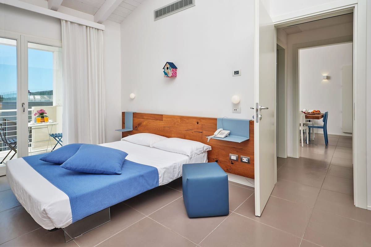 A modern hotel room with a bed, a small table with a chair, a balcony with a table and chairs, and a view of the sea.