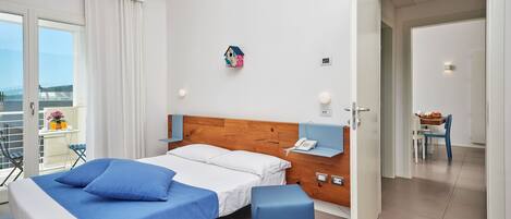 A modern hotel room with a bed, a small table with a chair, a balcony with a table and chairs, and a view of the sea.