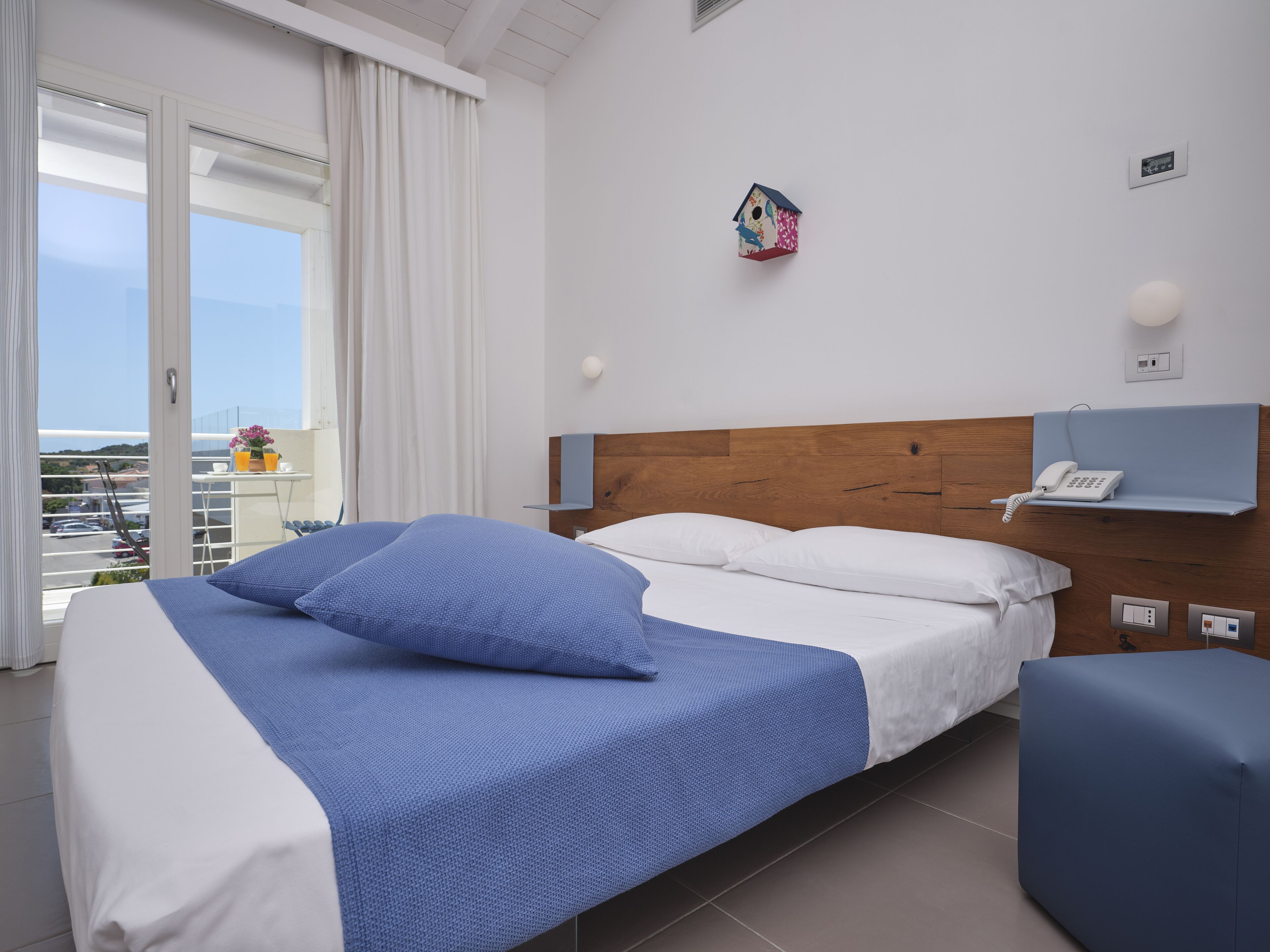 A hotel room with a bed, a bedside table, a phone, a wall-mounted light, and a view of the outside.