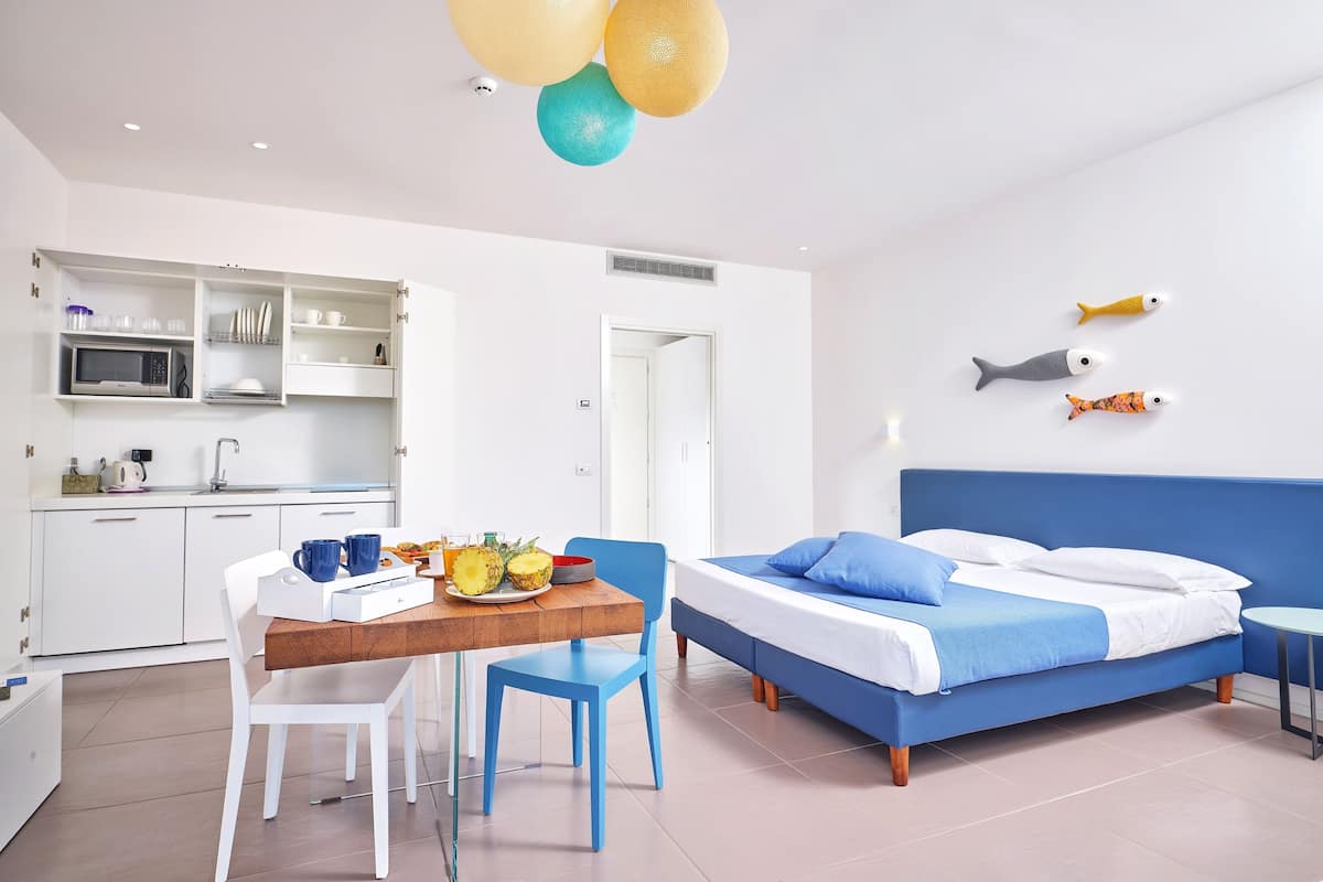 A modern hotel room with a kitchen area, a dining table with chairs, a bed with blue bedding, and decorative fish on the wall.