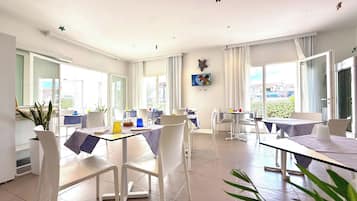 A bright, open dining area with white chairs and tables, large windows, and a city view.