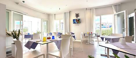 Un espace de salle à manger lumineux et ouvert, avec des chaises et des tables blanches, de grandes fenêtres et une vue sur la ville.