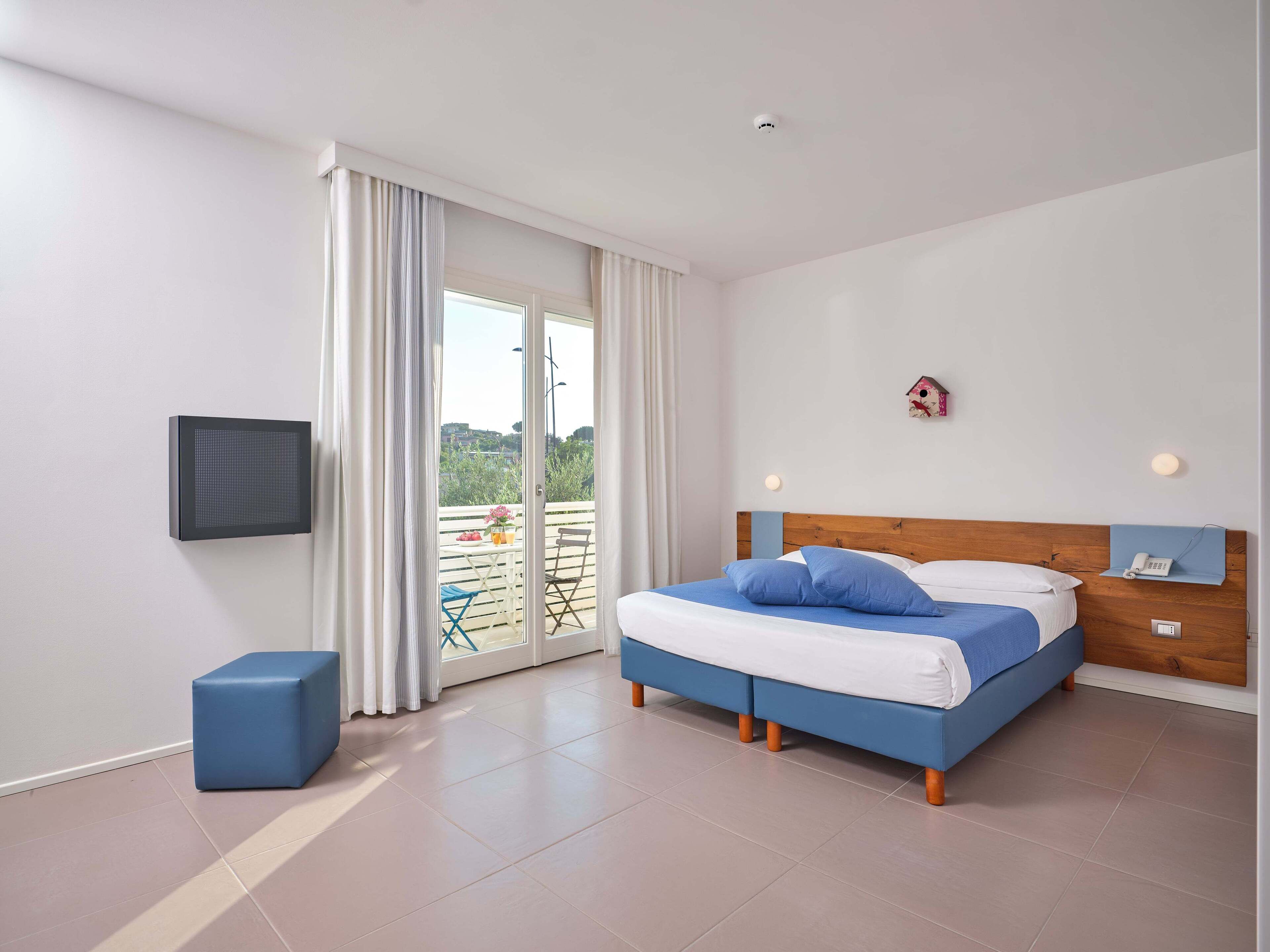 A modern bedroom with a large bed, a flat-screen TV mounted on the wall, and a balcony with a table and chairs.