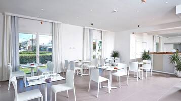 A modern dining area with white tables and chairs, large windows with curtains, and a view of the city.