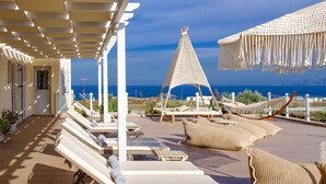 Un espacio lounge al aire libre iluminado por el sol, con sillones blancos, un dosel tejido y vistas al mar.