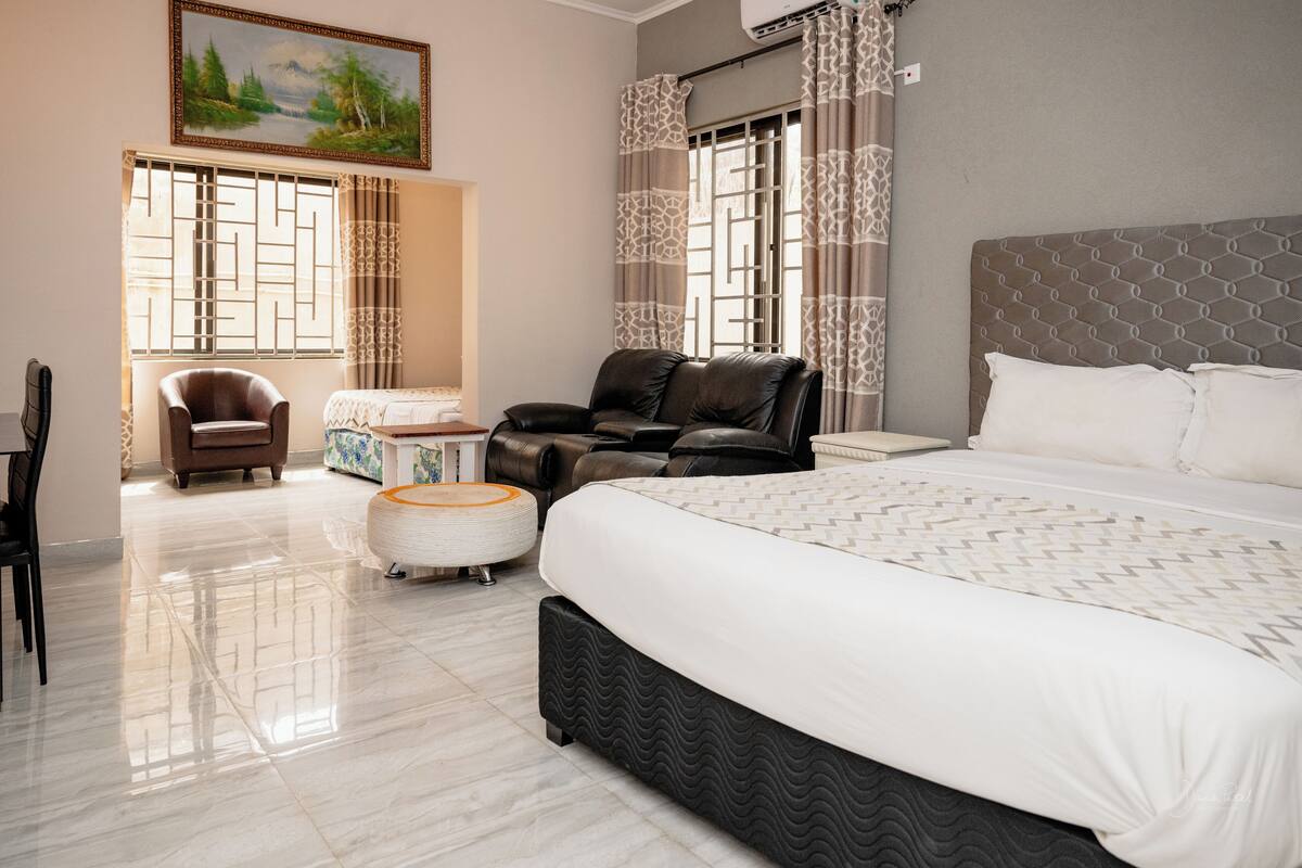 A modern hotel room with a large bed, two leather chairs, a small round ottoman, and a painting on the wall.