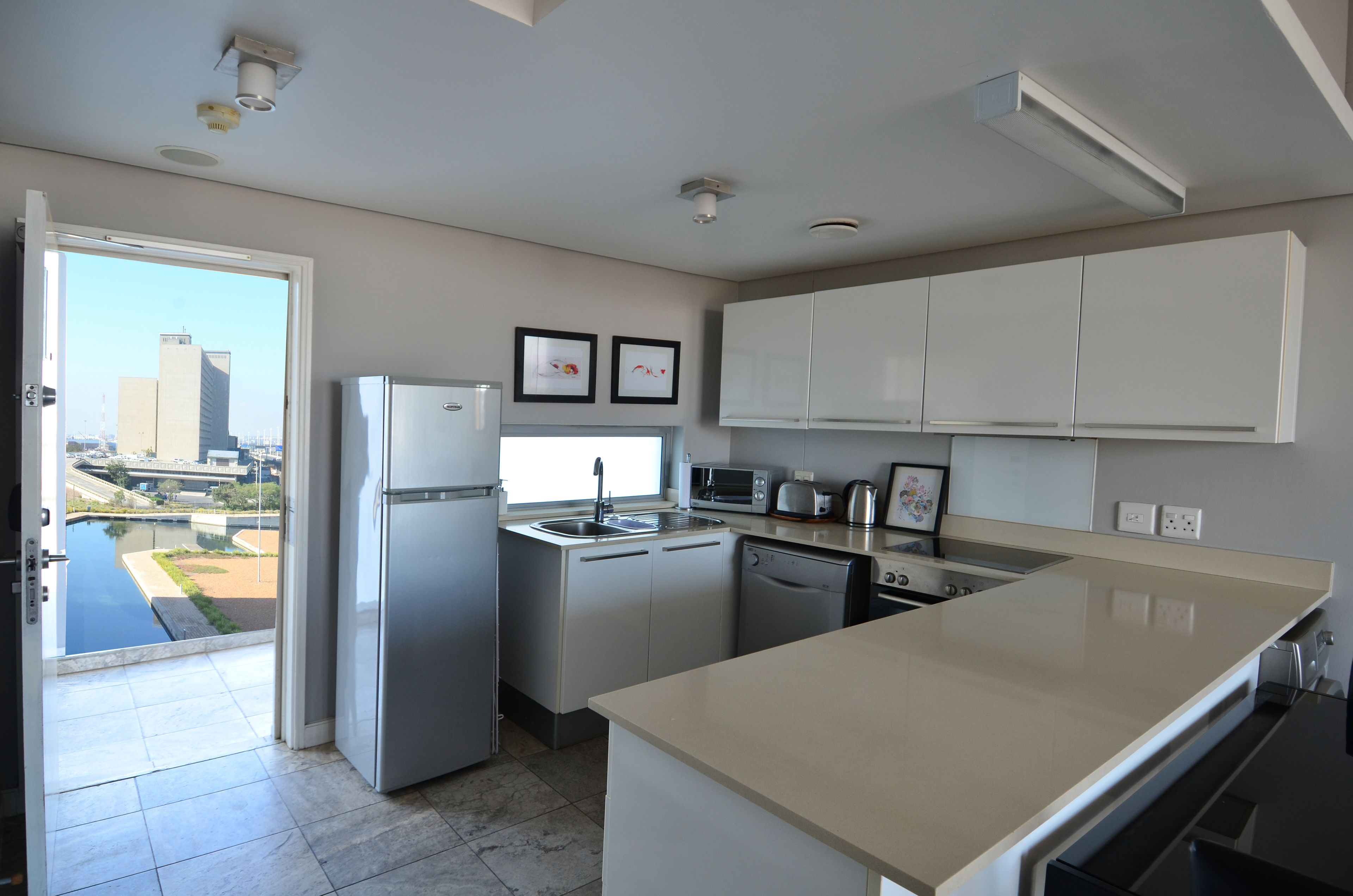 Cocina moderna con horno integrado, microondas y lavaplatos. Hay una isla grande con encimera color beige y vista a un paisaje urbano a través de una puerta abierta.