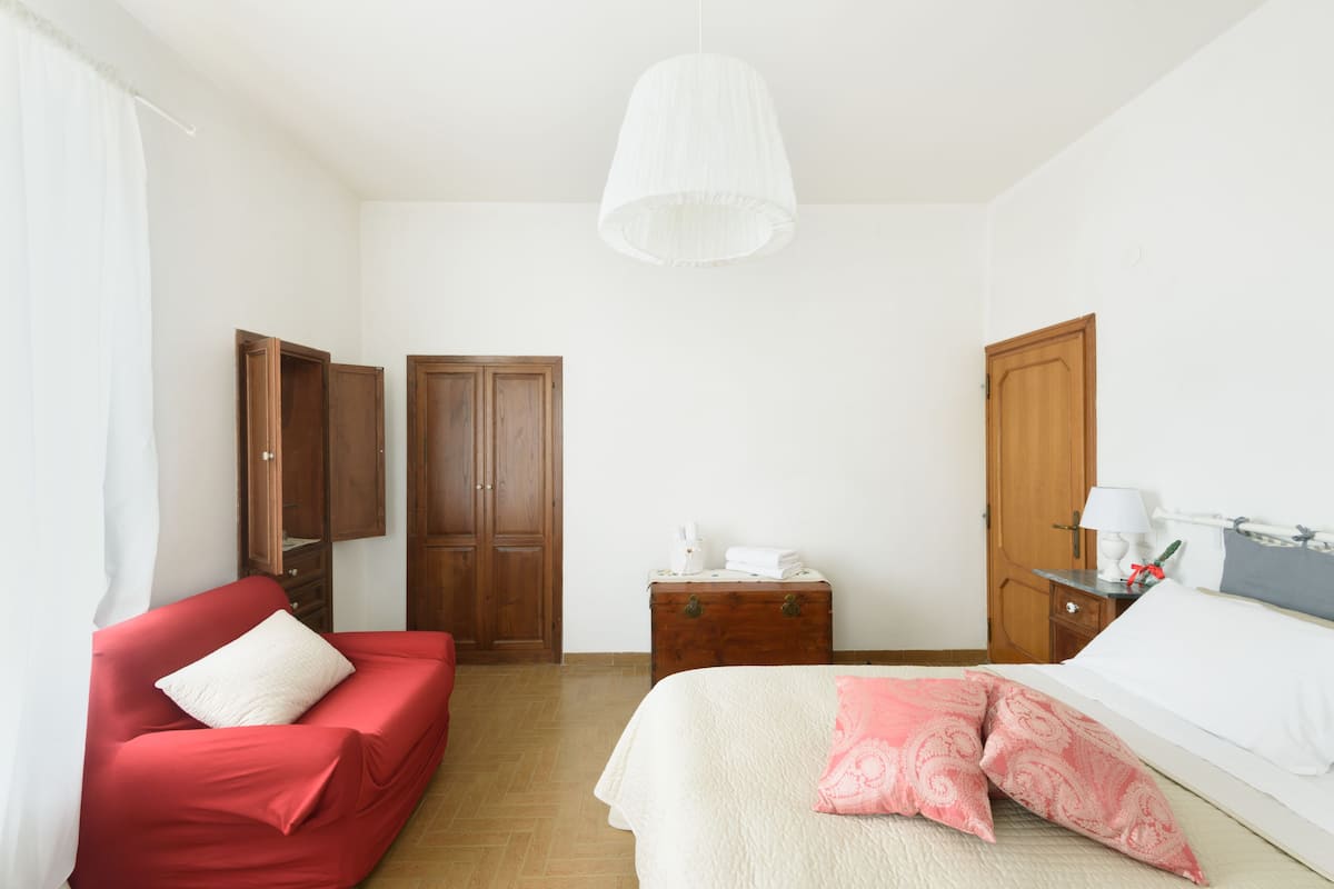 Ein Schlafzimmer mit einem roten Sofa, einem Bett mit weißen und rosafarbenen Kissen, einem Holzschrank, einem Nachttisch mit Lampe und einer Pendelleuchte.
