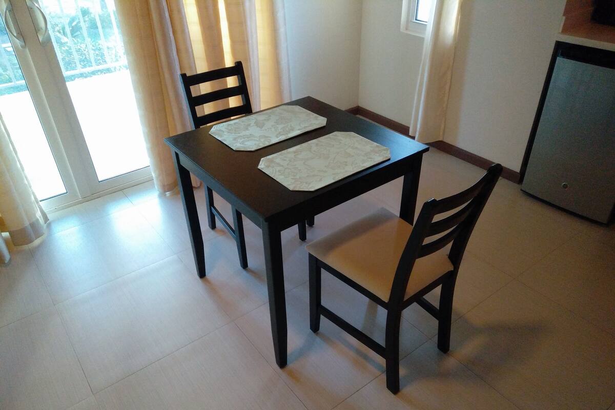 Un comedor con una mesa, cuatro sillas, un refrigerador pequeño y una ventana con cortinas.