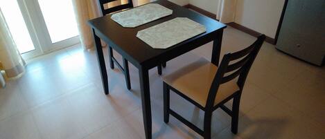 Un comedor con una mesa, cuatro sillas, un refrigerador pequeño y una ventana con cortinas.