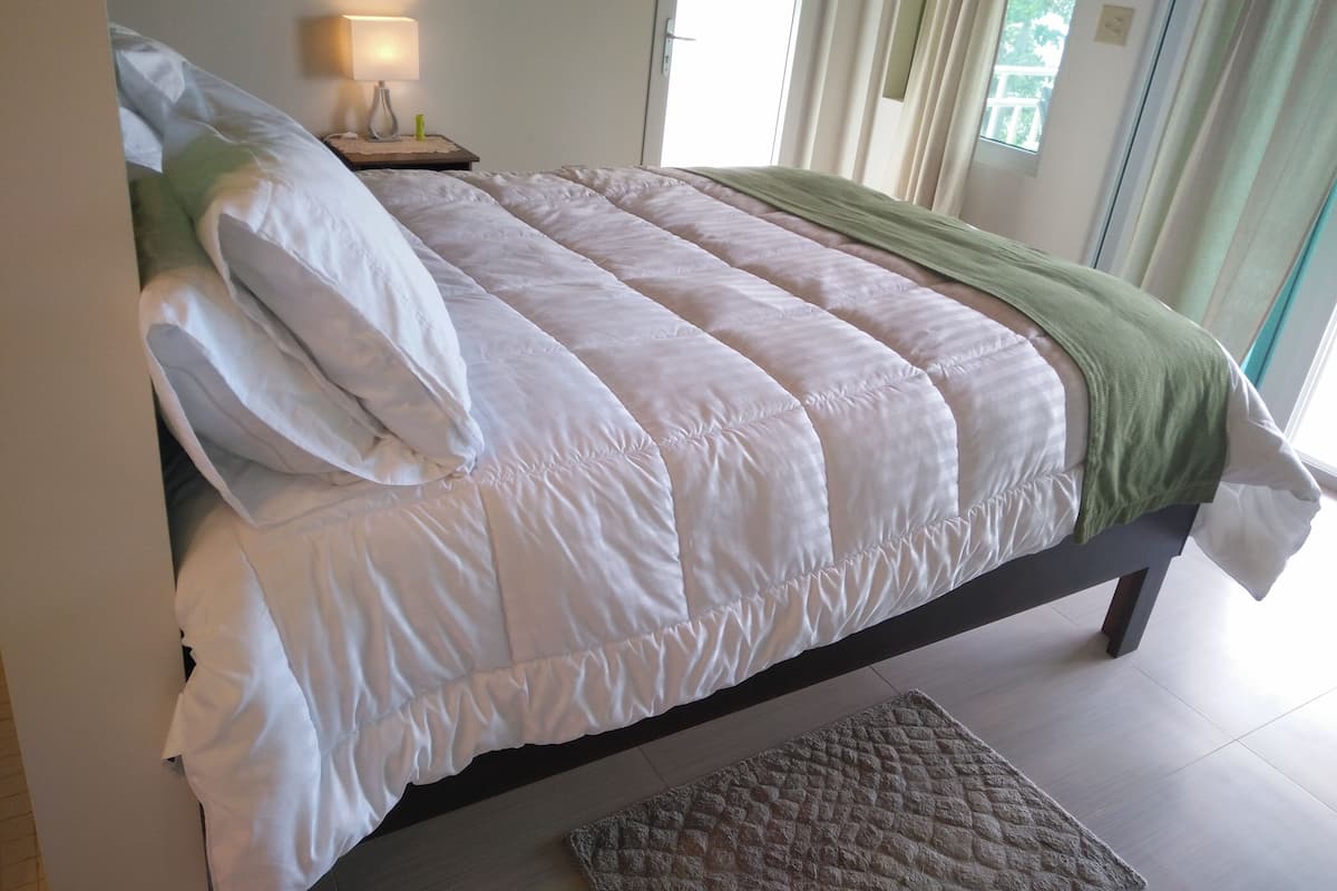 Una cama bien hecha con ropa de cama blanca y una manta verde, una lámpara de noche y vistas al exterior a través de una ventana con cortinas.