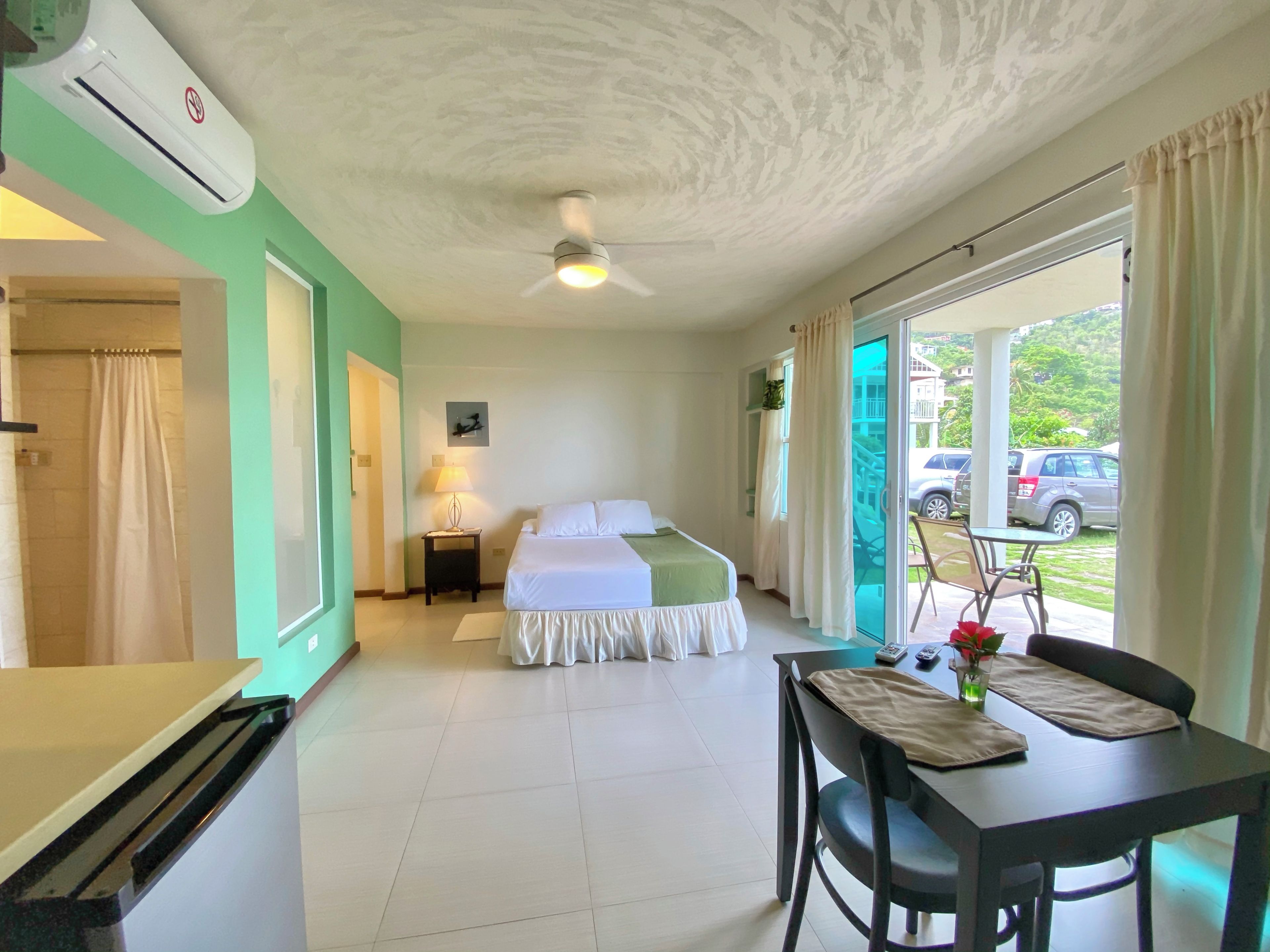 Habitación de hotel con cama, escritorio, silla, mesa, aire acondicionado y ventilador de techo, con vistas a coches aparcados fuera.