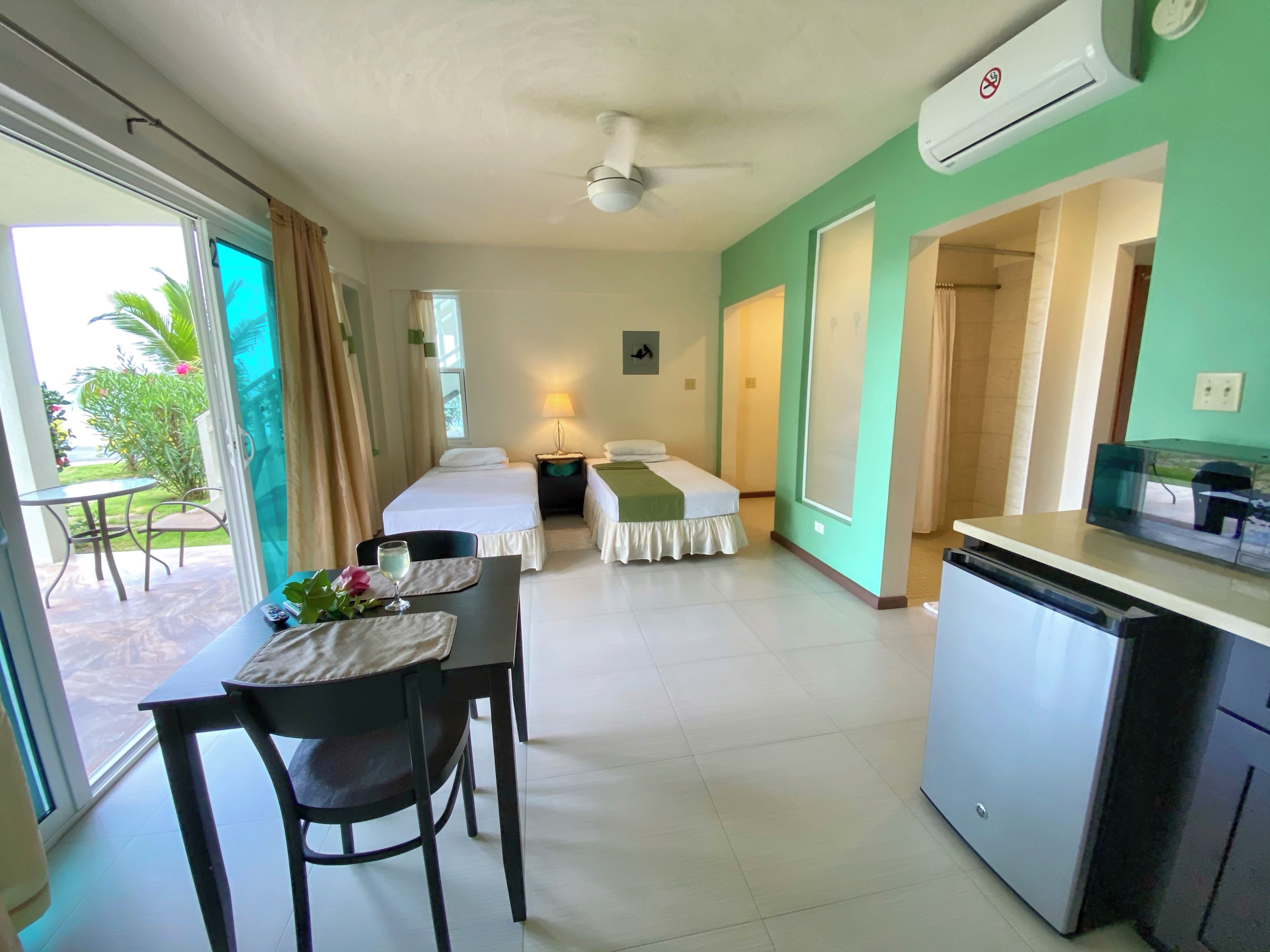 A hotel room with a bed, a desk with a chair, a table with a glass of water, and a kitchenette.
