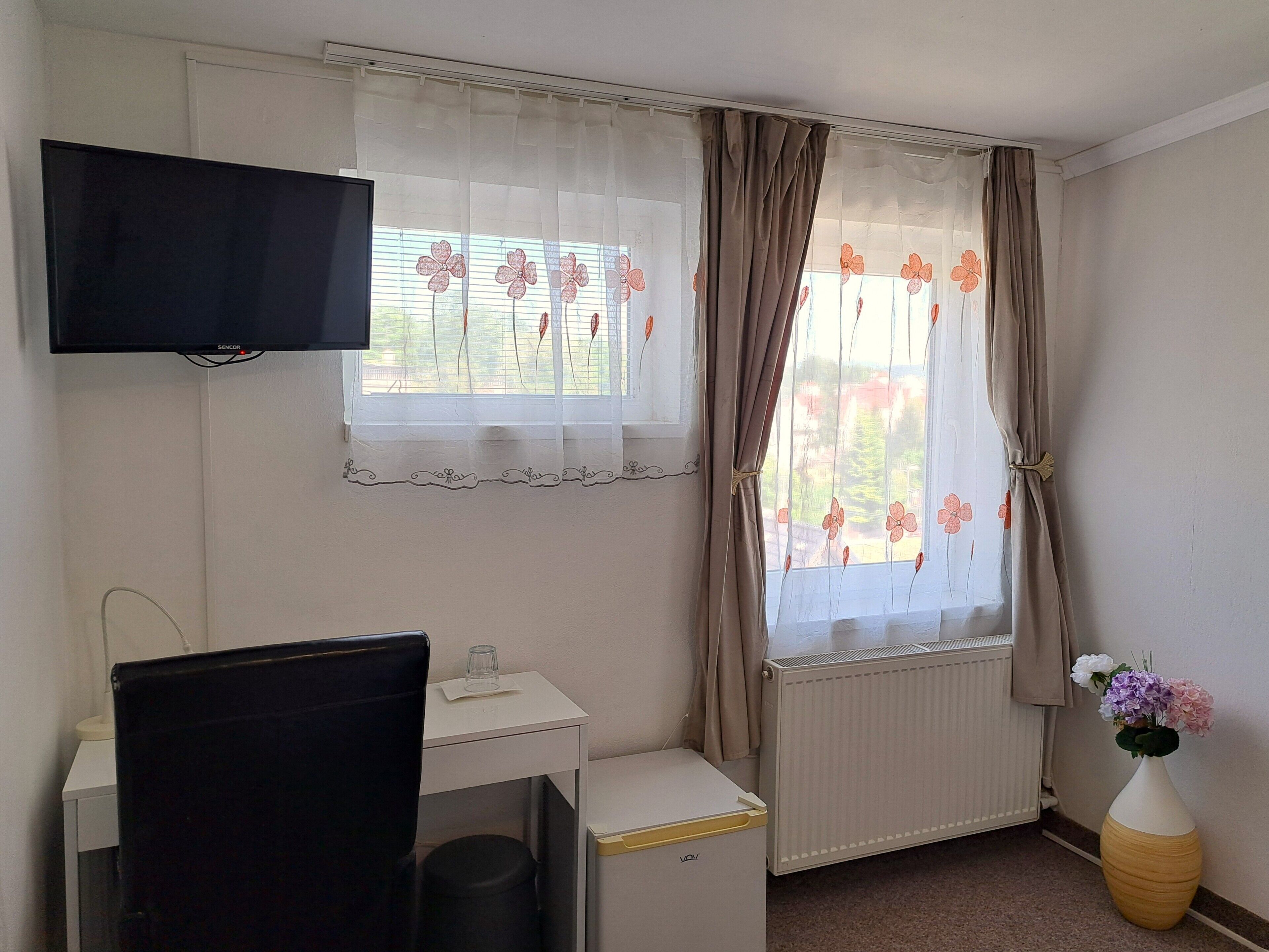 Ein Zimmer mit Schreibtisch, Stuhl, Wandfernseher, Heizkörper und einem fenster mit Vorhängen.