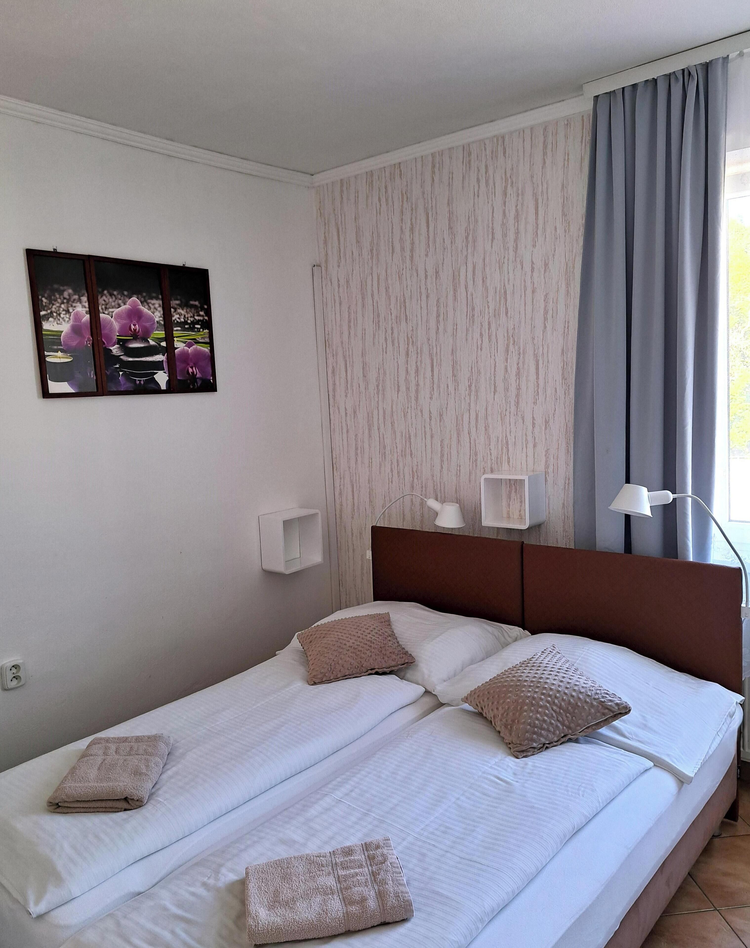 Un letto rifatto con cuscini e un asciugamano piegato, un quadro appeso al muro, una lampada da comodino e una finestra con tende.