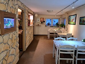 Een kamer met stenen muren, een eettafel met stoelen, een klok en een raam met planten.