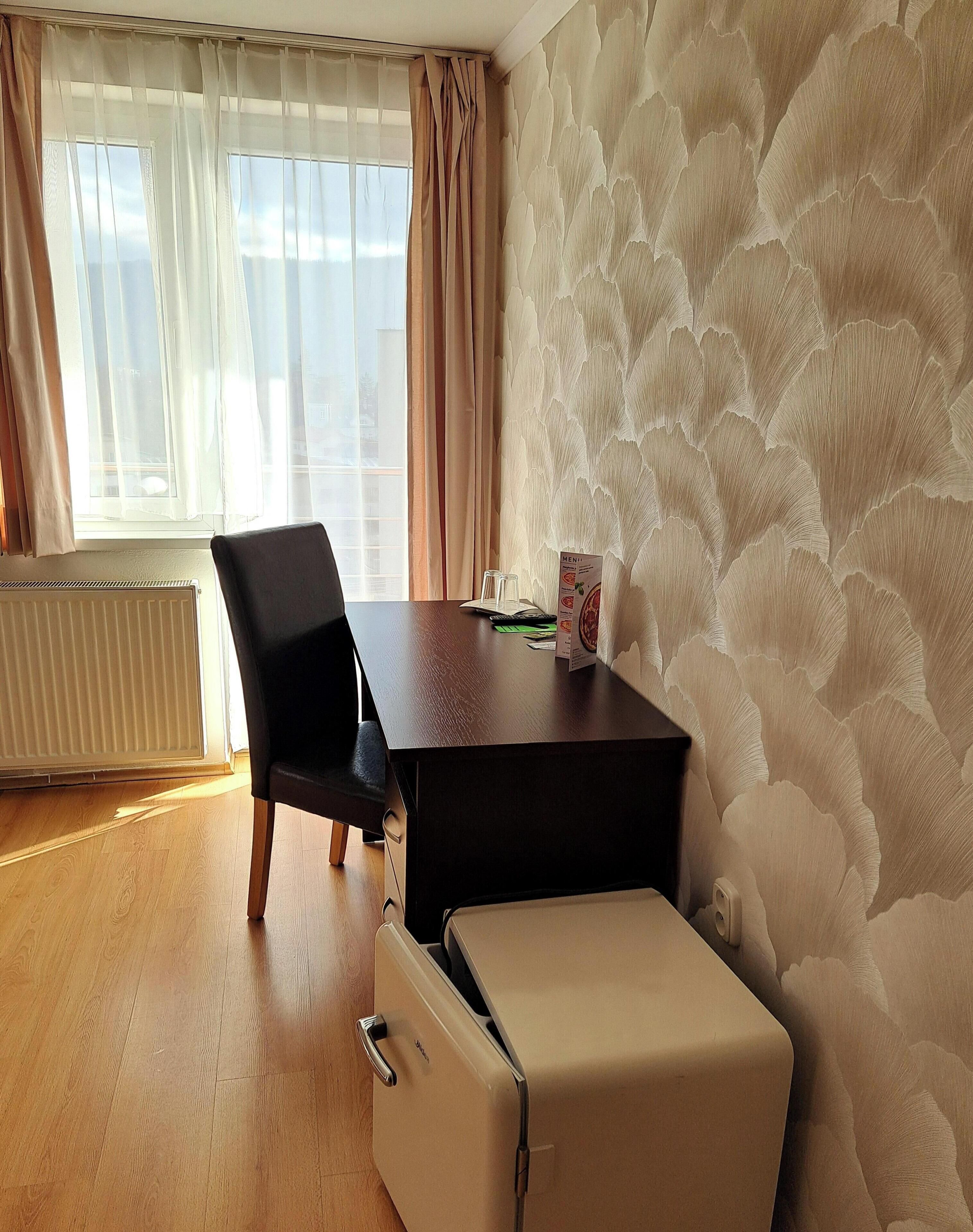 Une pièce avec un plancher en bois, une chaise noire, une petite table avec un menu, un radiateur et une fenêtre avec des rideaux.