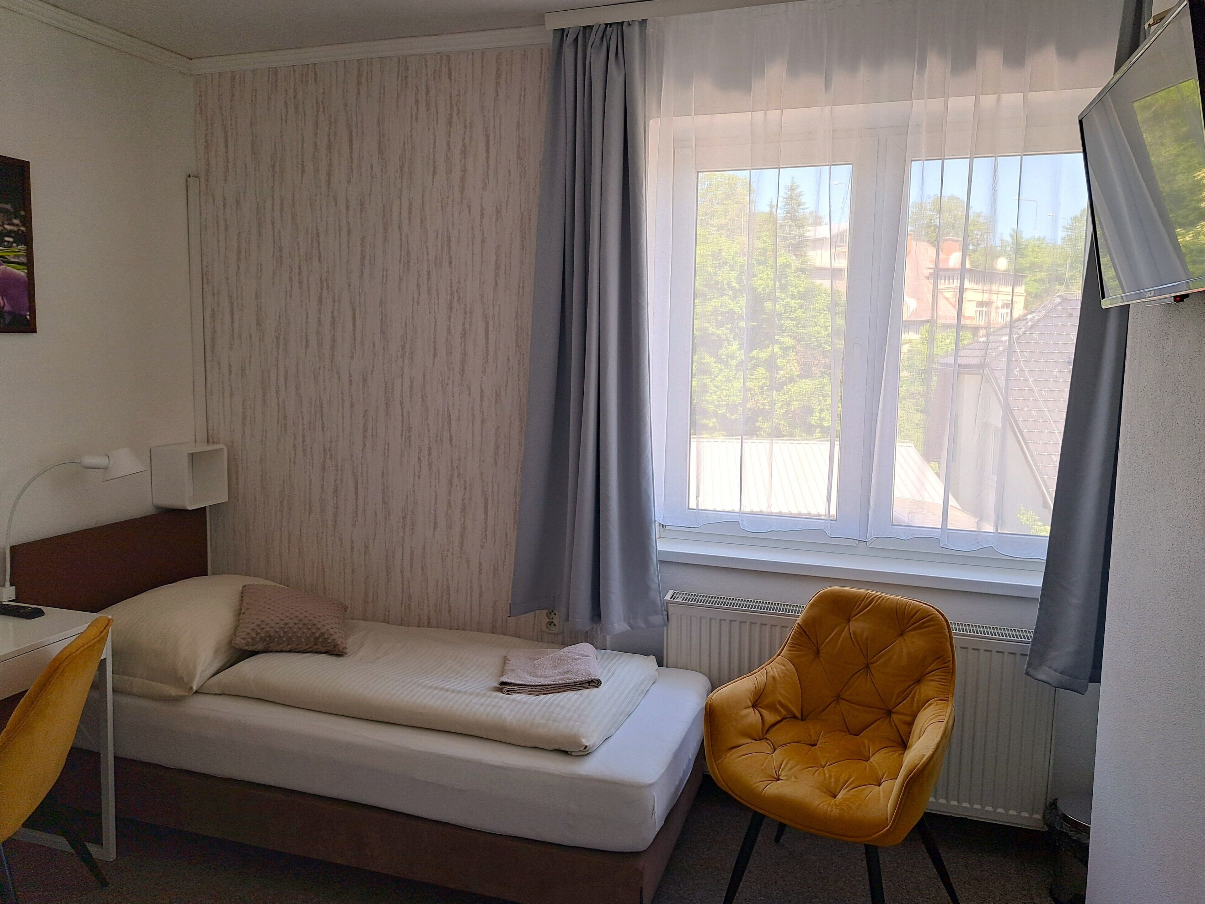 Un lit simple avec une tête de lit, une table de chevet avec une lampe, une chaise et une fenêtre avec des rideaux.