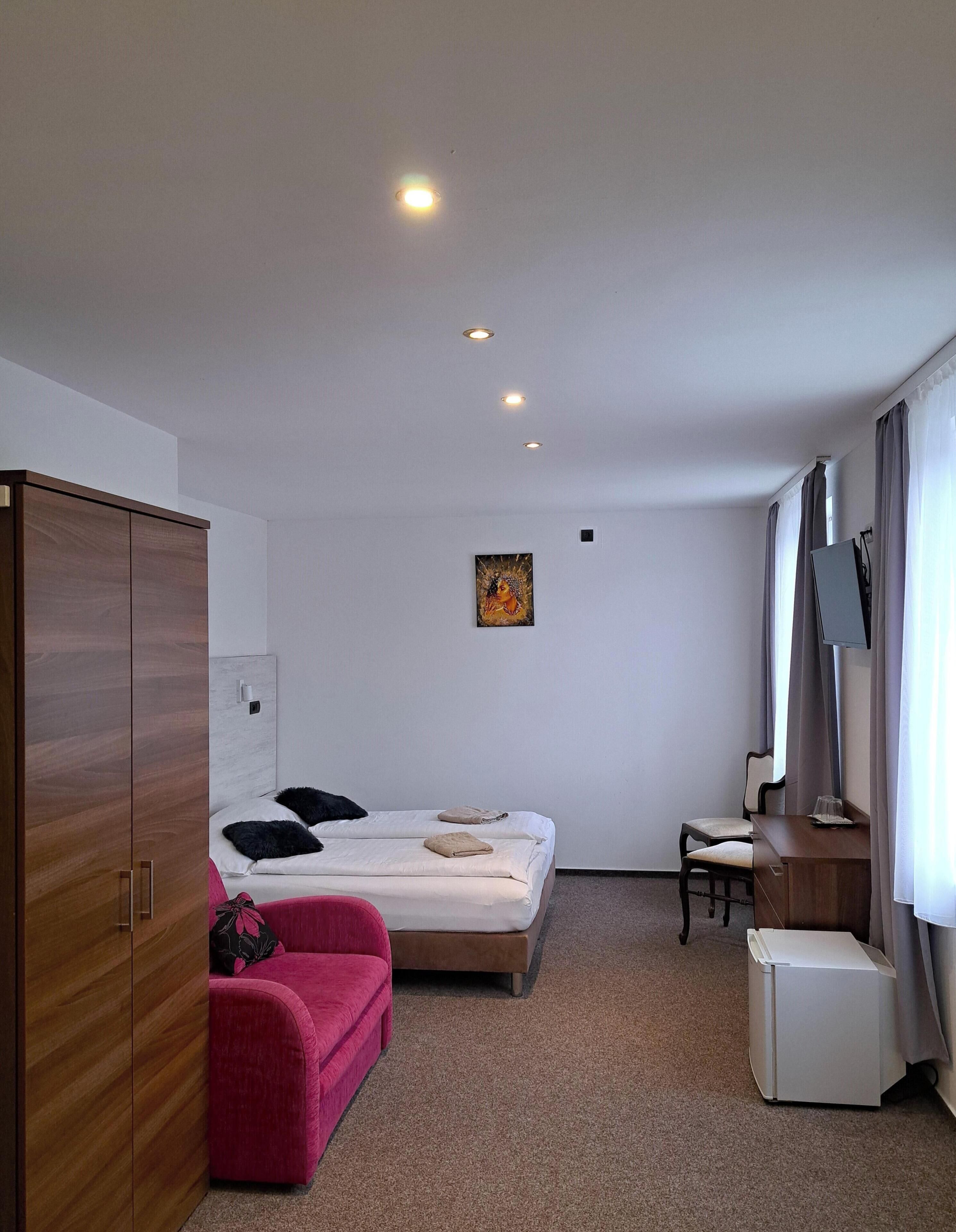 A hotel room with a bed, a desk, a wardrobe, a pink chair, and a painting on the wall.
