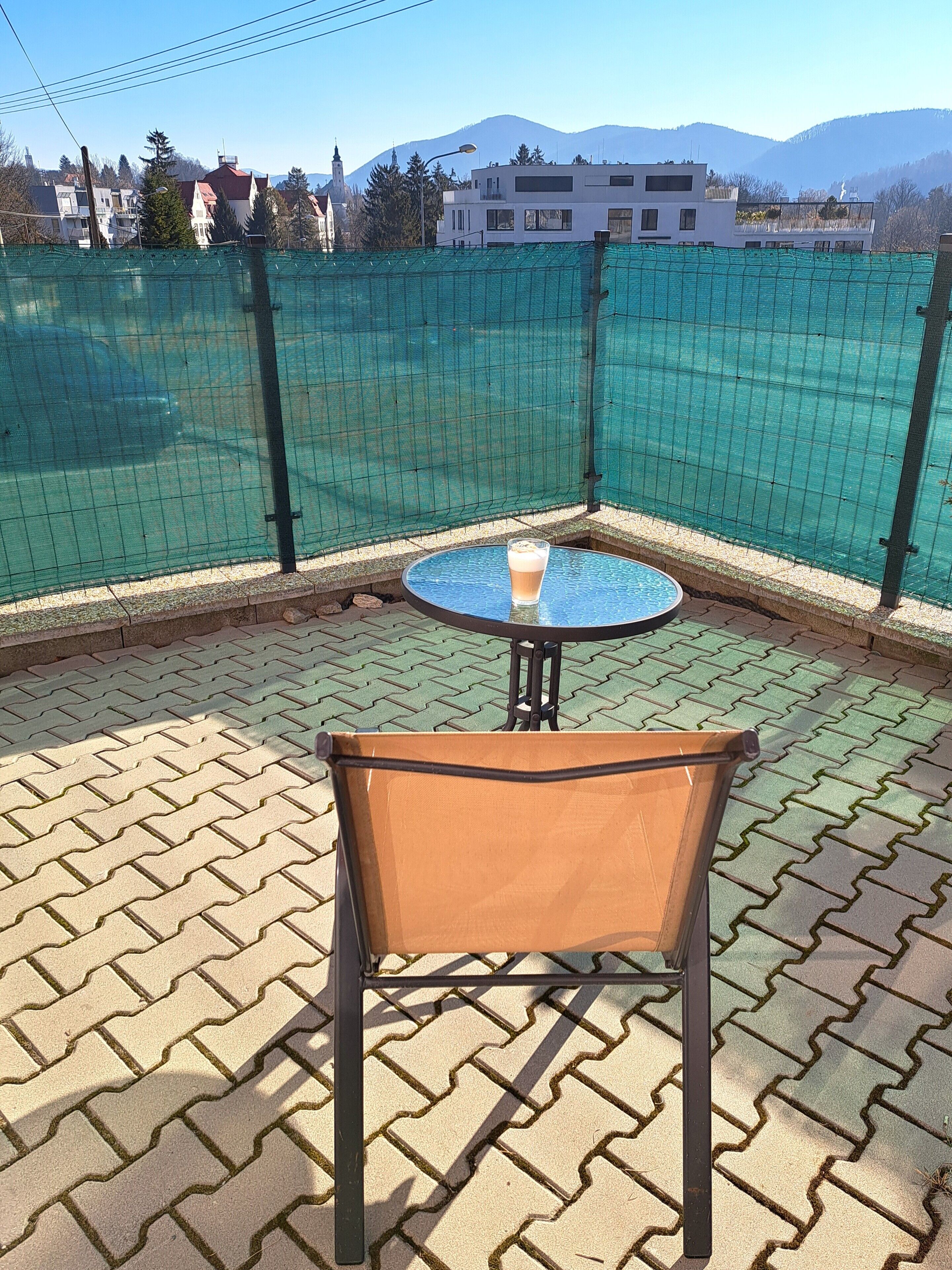 A corner patio with a table and chair, a green netted enclosure, and a view of buildings and mountains.