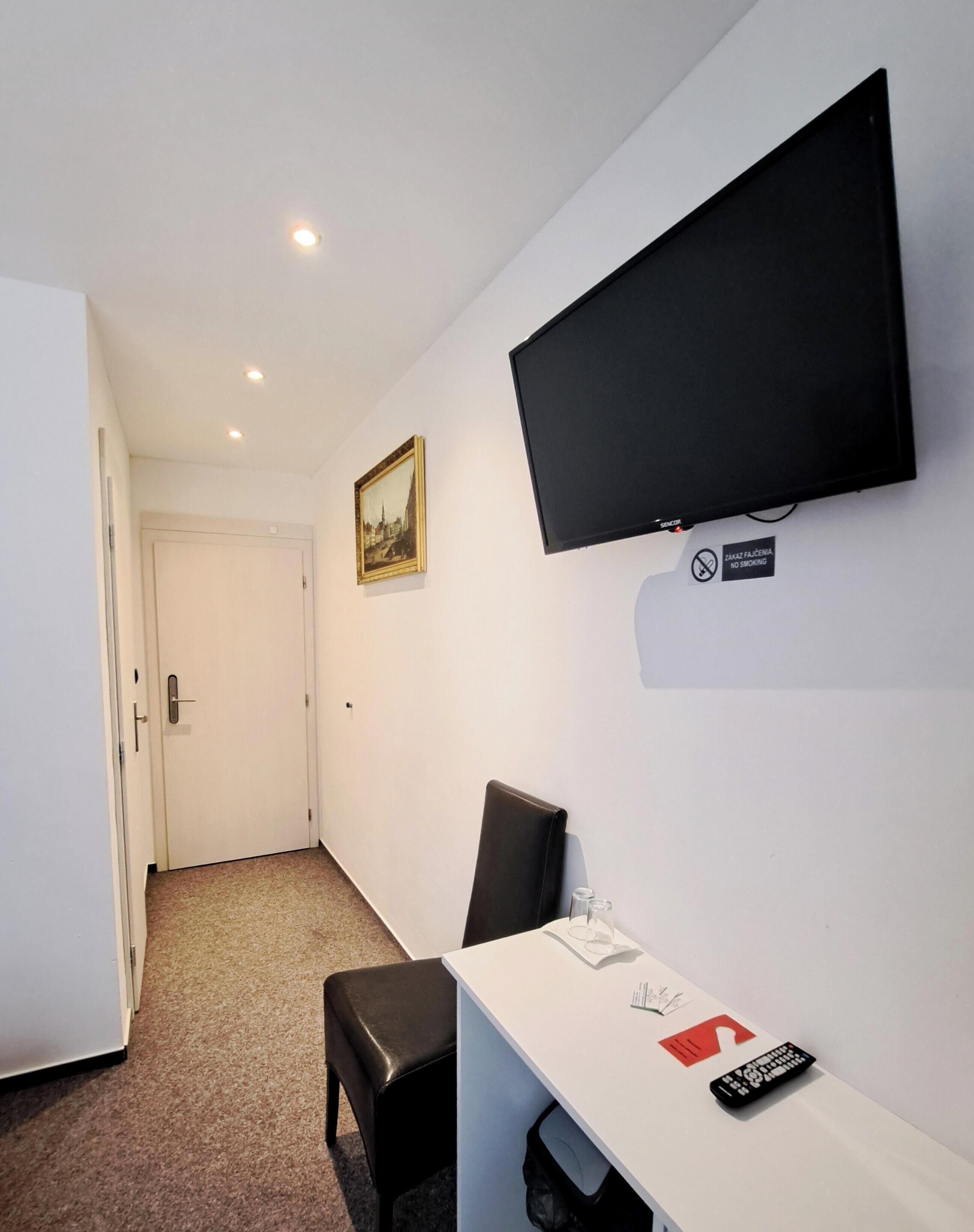 Una camera d'albergo con un letto, una scrivania con una sedia, una televisione a muro, una porta e un pavimento rivestito di moquette.