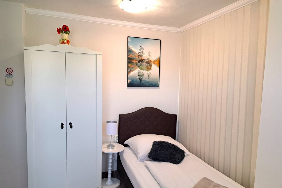 Et soveværelse med seng, garderobeskab, natbord, lampe, et maleri og et vindue med persienner.