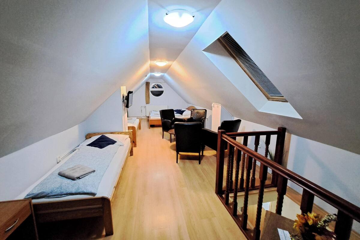 Et aflangt rum på loftet med skråvægge, en trætrap og et lille siddeområde med en stol og et bord.