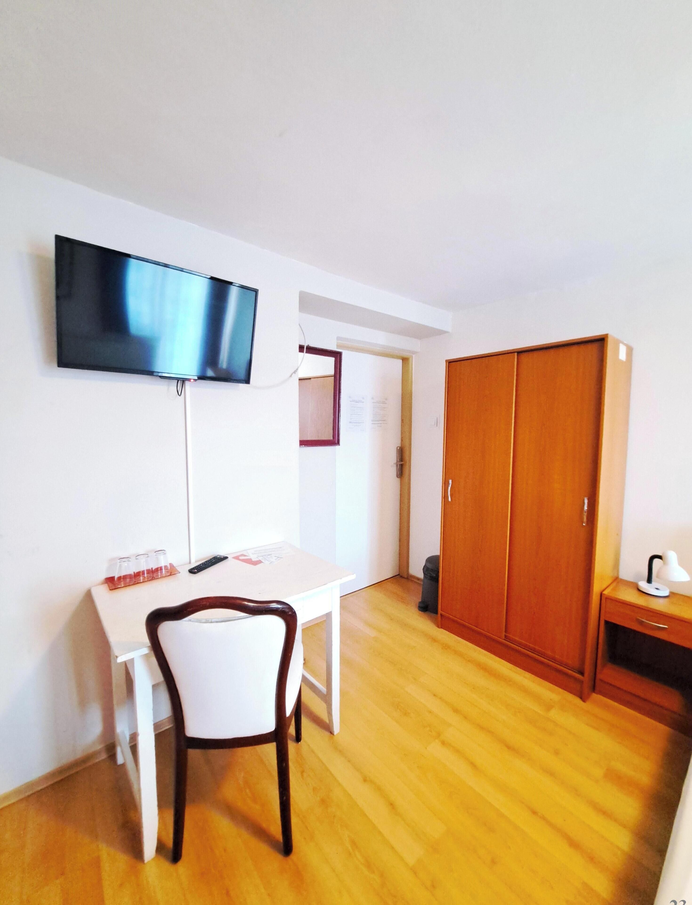 A room with a TV mounted on the wall, a wooden wardrobe, a white table with a chair, and a trash bin.