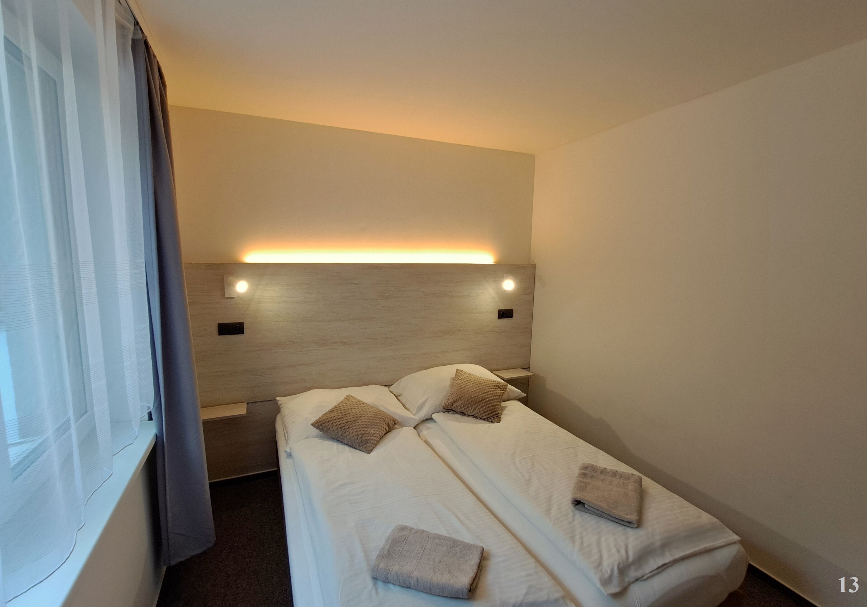 Une chambre d’hôtel avec deux lits, une tête de lit dotée d’un éclairage intégré, une fenêtre avec des rideaux et une petite table de chevet.